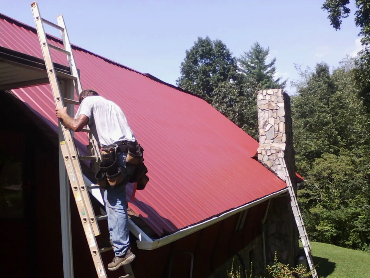 Roofer on a ladder beside a red metal roof and stone chimney — Soffit & Fascia Repair by Roofing Brothers in Knob Lick