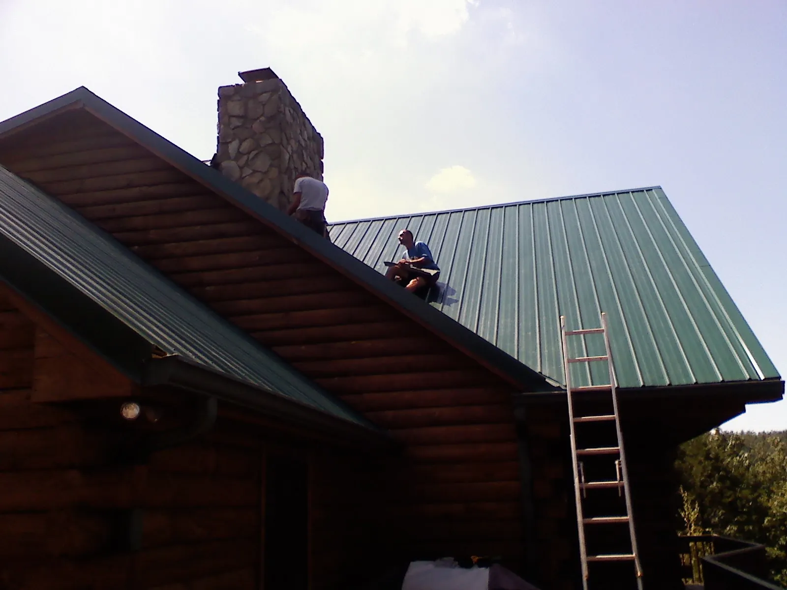 Roofers installing green metal panels beside a stone chimney on a log home in Knob Lick